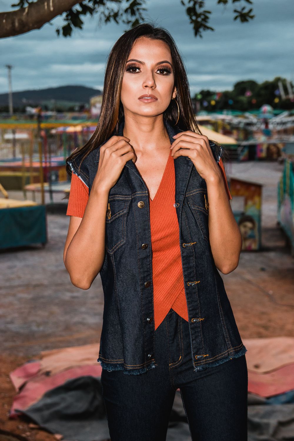 Confident woman posing outdoors in fashionable denim vest and orange top with a fairground background.