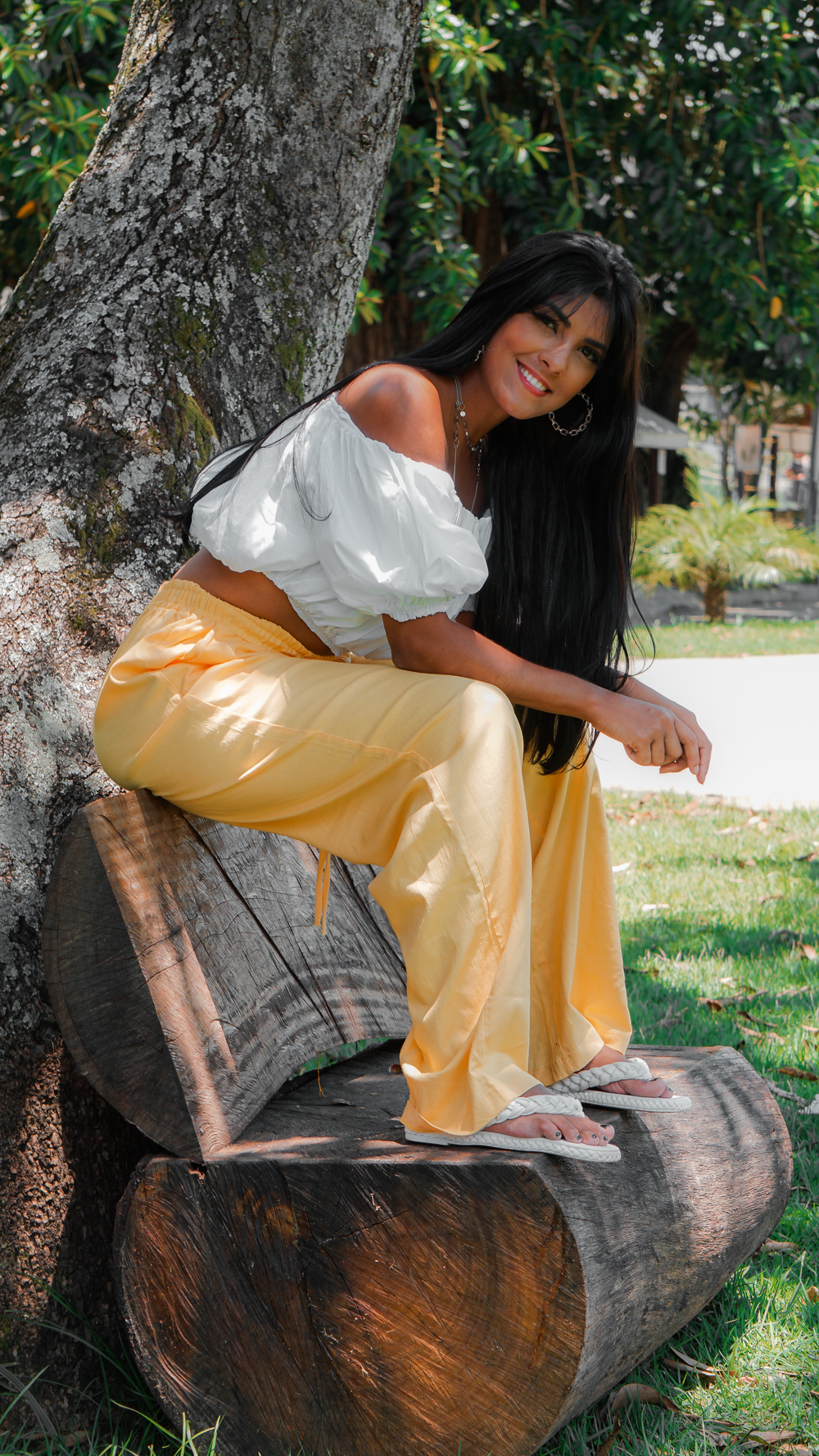 A fashionable woman wearing a crop top and orange pants sitting on a bench in a sunny park.