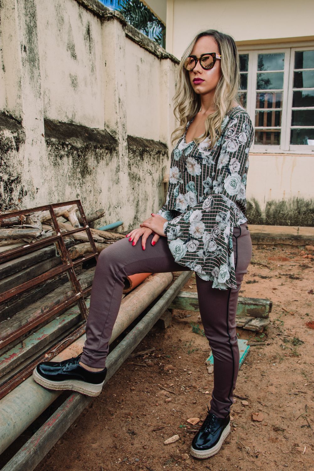 Stylish woman in patterned blouse and sunglasses posing by abandoned building.
