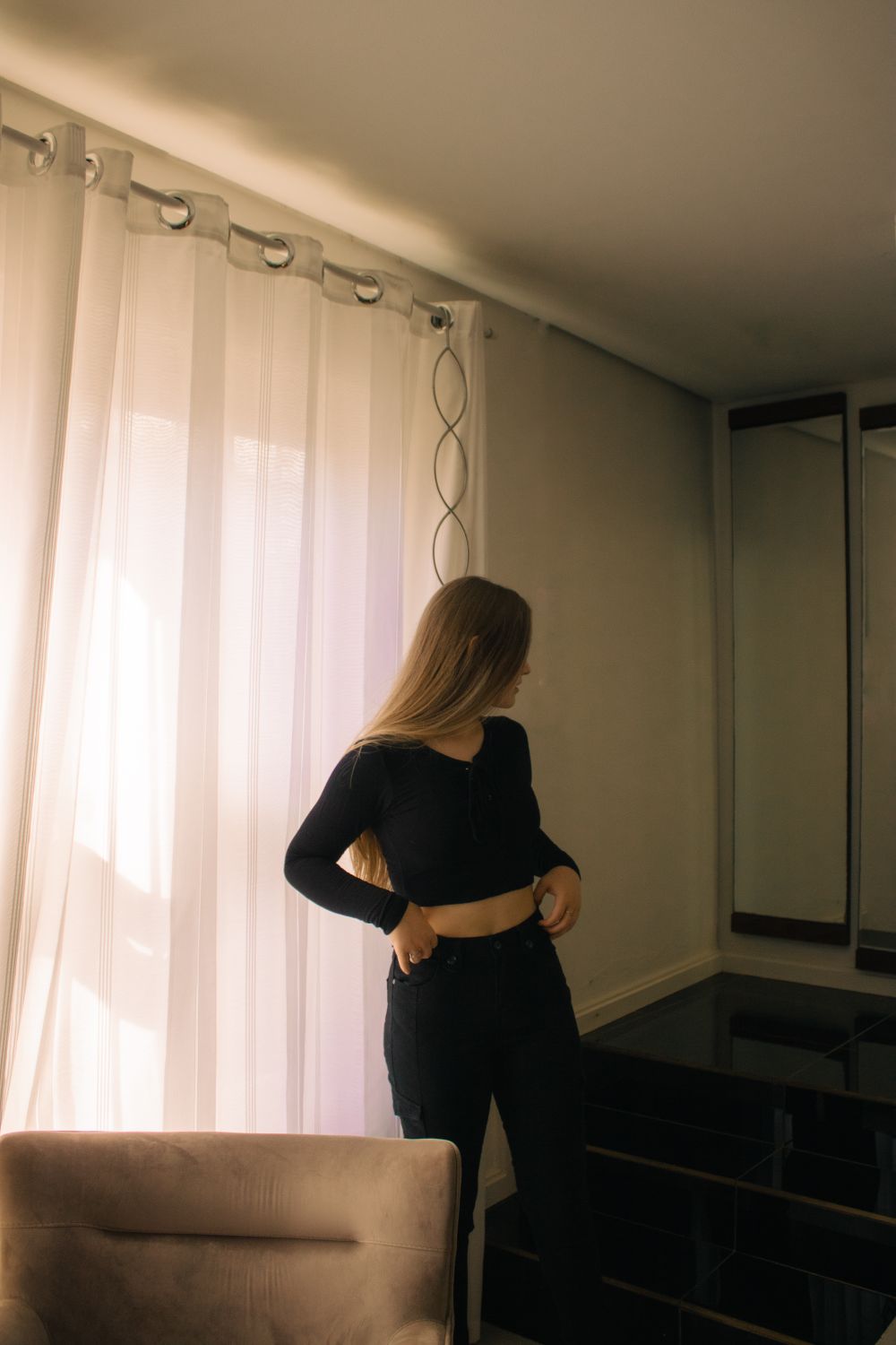 Woman standing indoors wearing black outfit near window with sheer curtains.