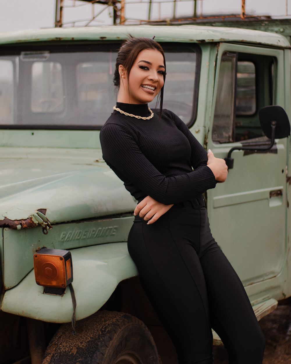 A woman in black attire smiling while leaning against a vintage green truck outdoors.