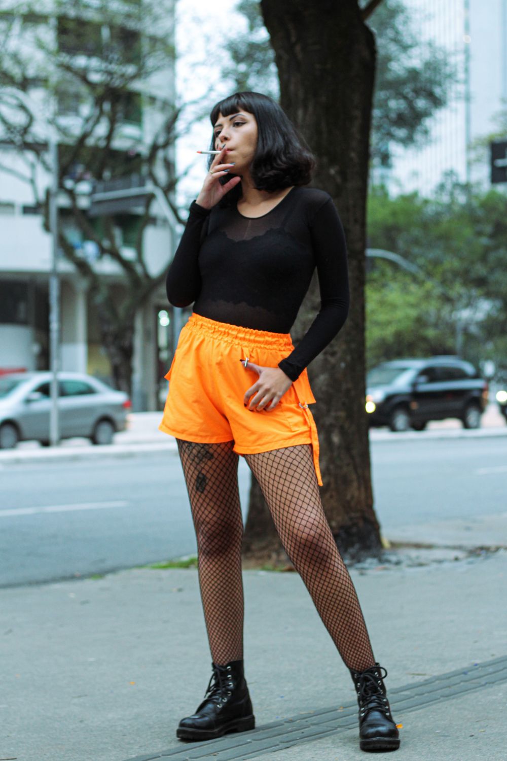 A fashionable woman stands confidently on a city sidewalk while smoking a cigarette.