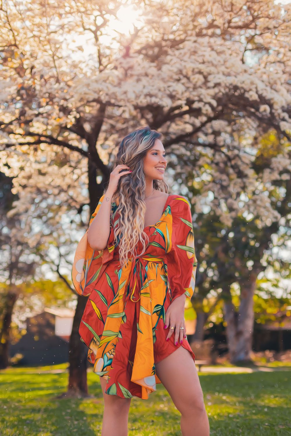 A smiling woman with curly hair in a vibrant floral dress enjoys a sunny day in a Bauru park.