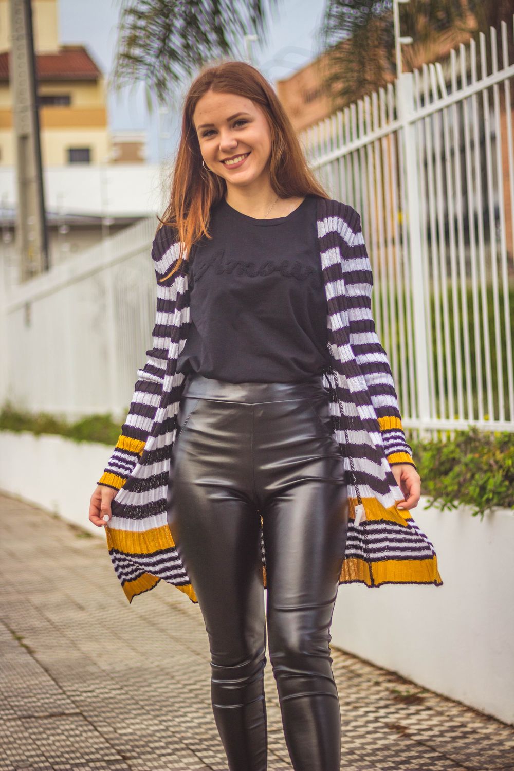 A happy woman in striped cardigan and leather pants posing outdoors with a bright smile.