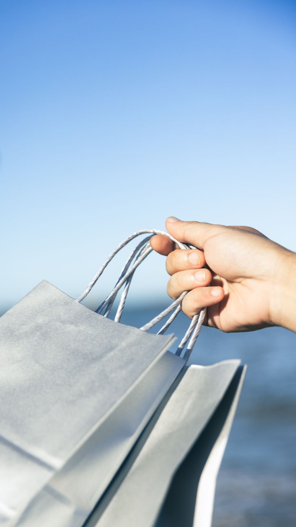 Close-up of a hand holding shopping bags by the beach, evoking leisure and travel vibes.