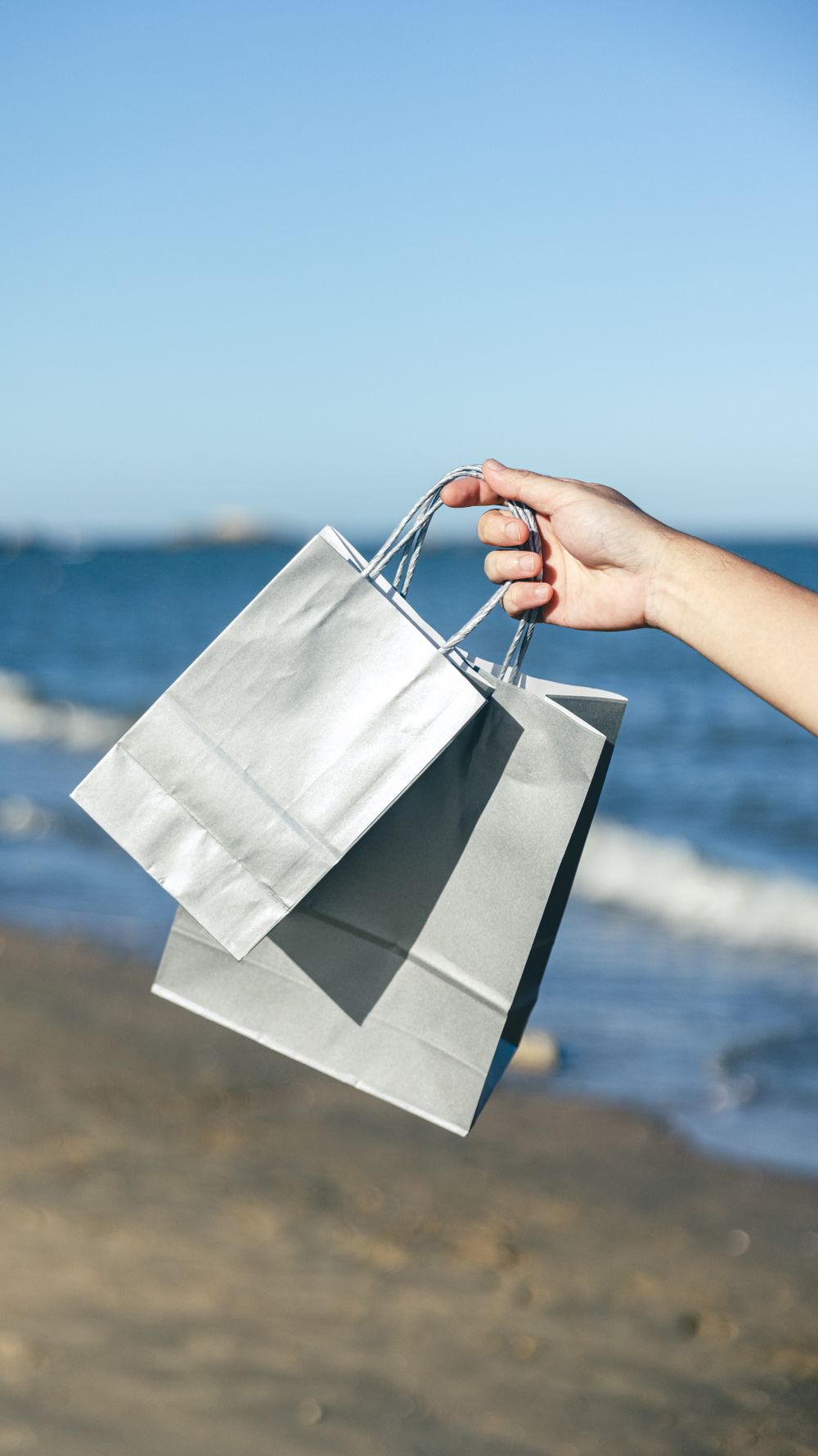 A close-up of a hand holding shopping bags on a sunny beach day.