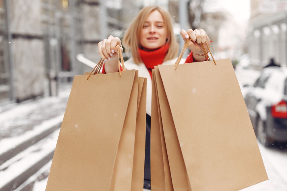 Woman smiling and holding paper bags on a snowy street, outdoor shopping scene.