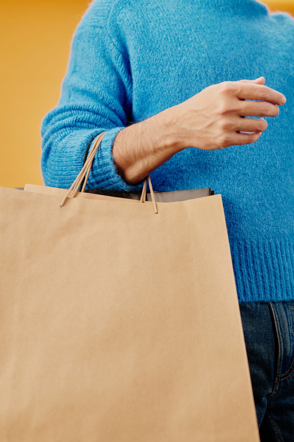 A person in a blue sweater holding a large paper shopping bag against a yellow background.