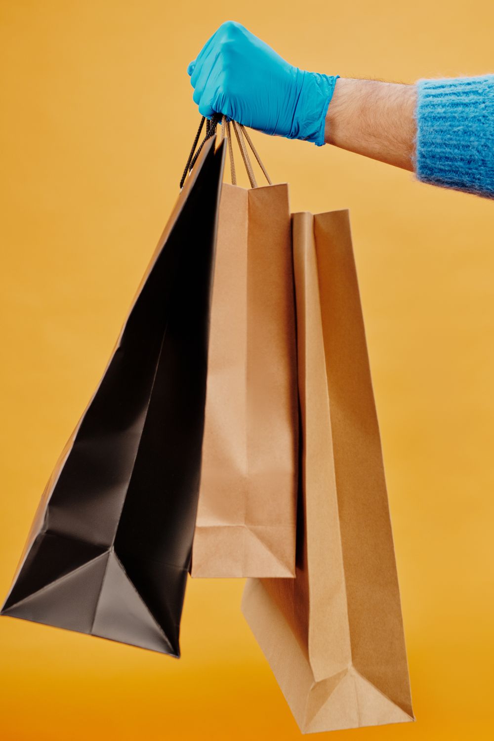 A hand in a blue glove holds paper shopping bags against a vibrant yellow background.