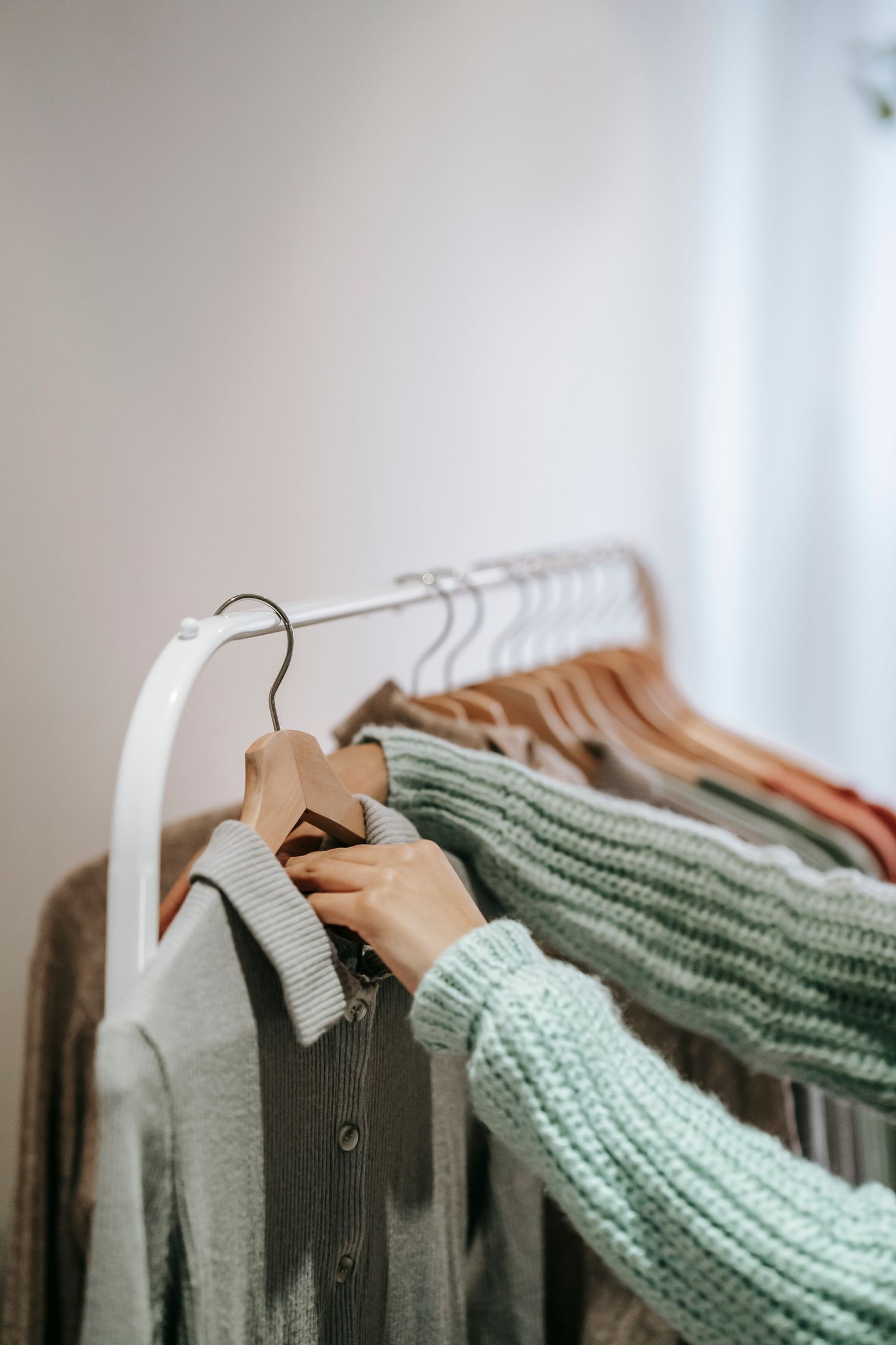 Crop unrecognizable female in knitted wear touching wooden hanger with clothes on stand in building