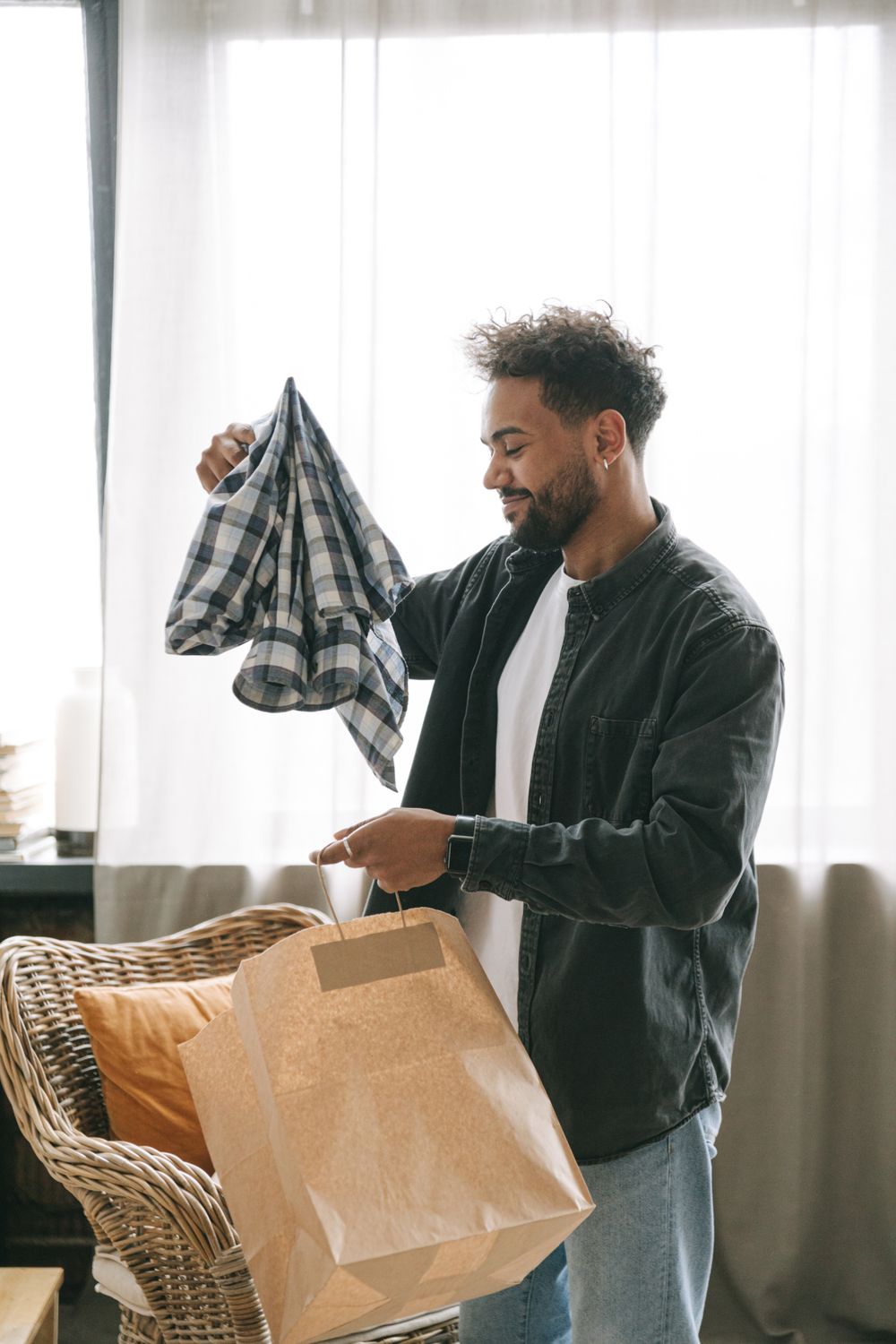 Young man organizing clothing indoors, holding a checkered shirt above a paper bag.