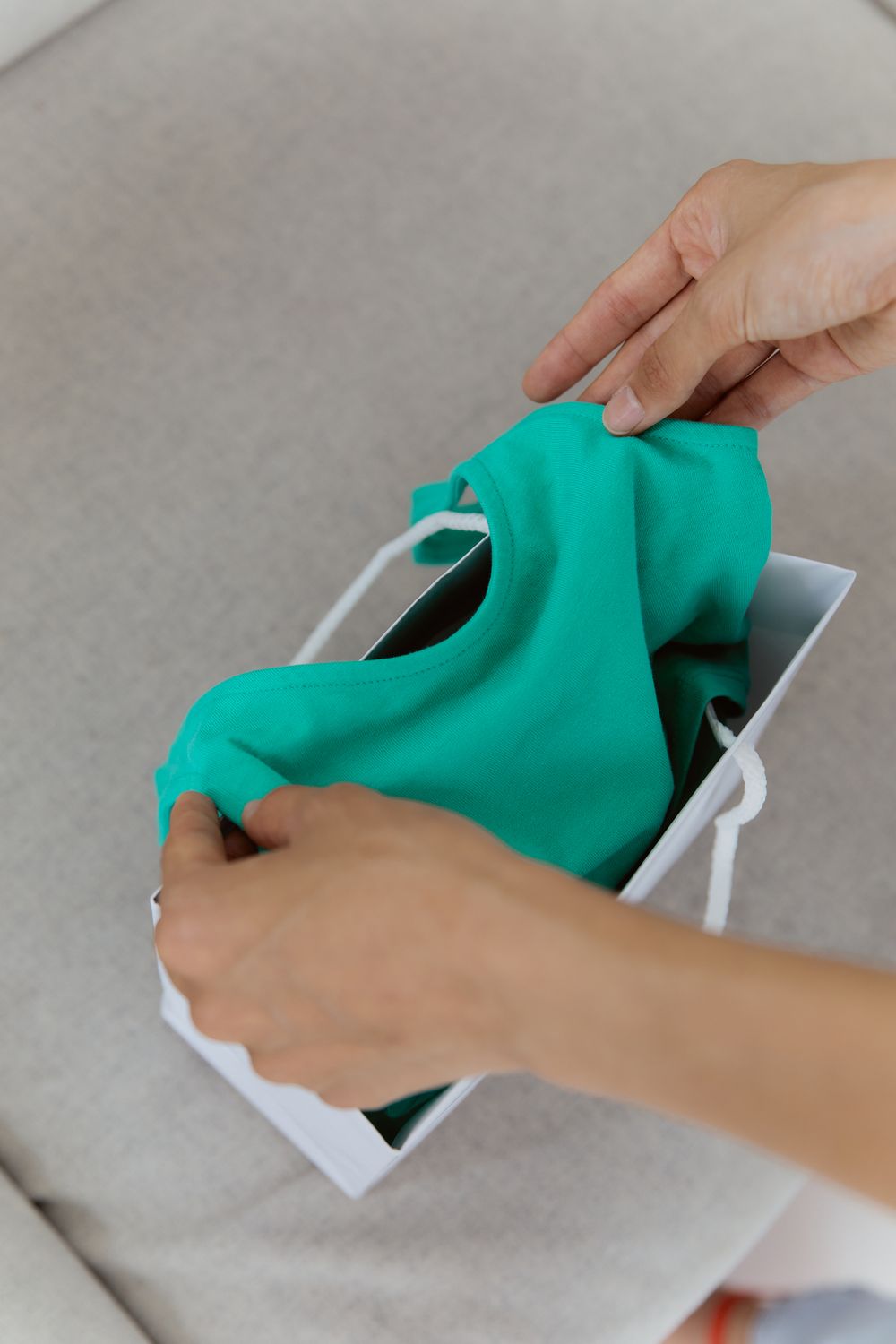 Close-up of hands unpacking a green shirt from a white shopping bag indoors.