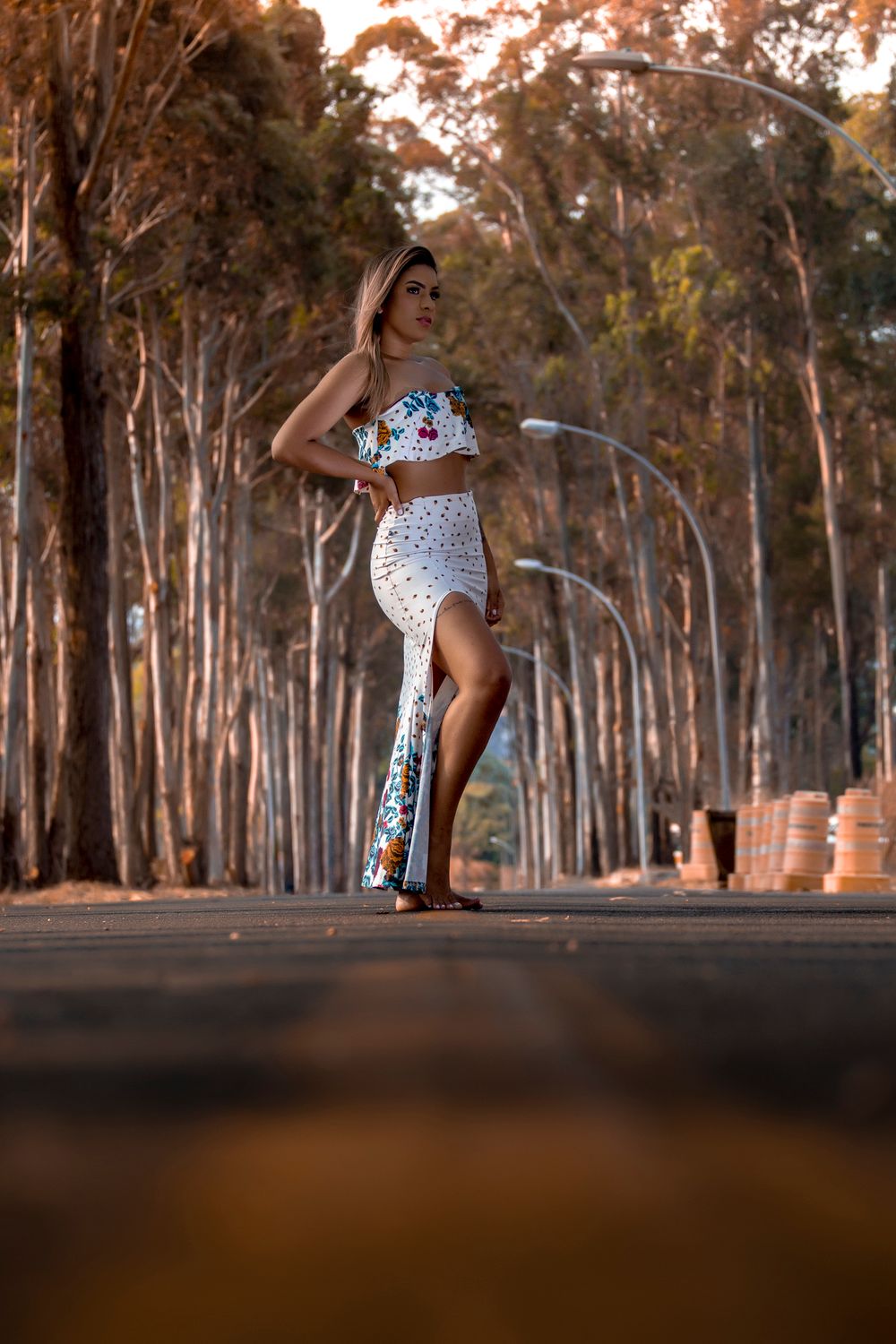 A fashionable woman in a floral outfit poses on a sunlit road lined with tall trees, showcasing a summer style.