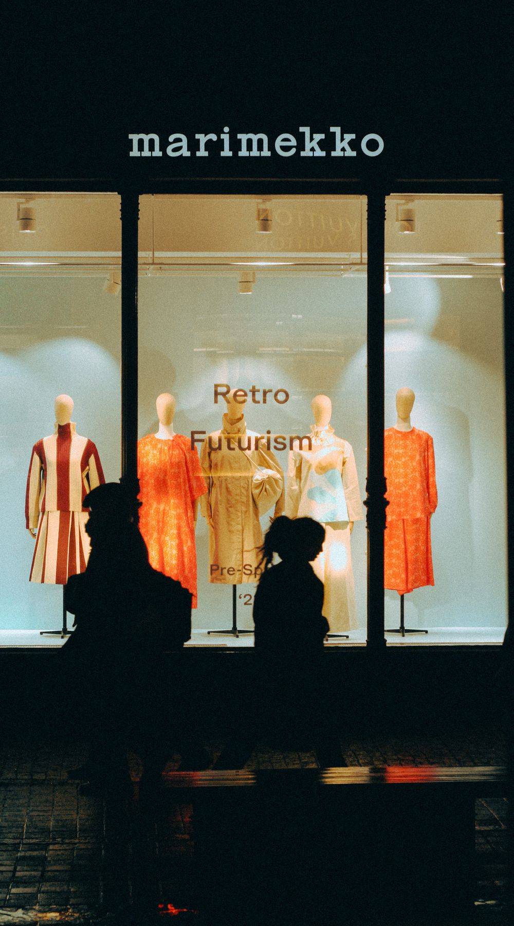 Silhouettes of people walking past a chic storefront display at night, featuring vibrant fashion.
